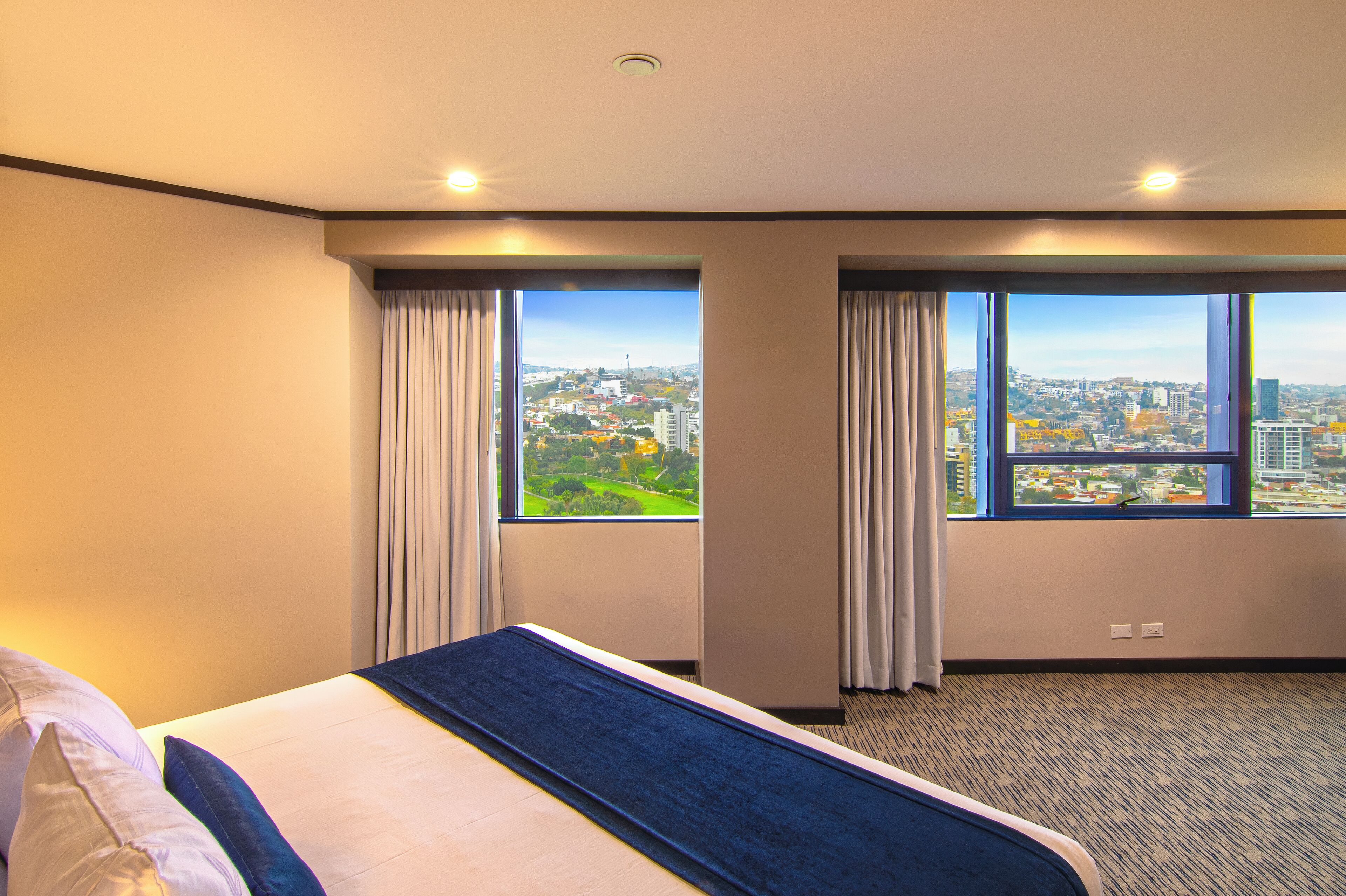 Quarto de hotel com uma cama grande, duas janelas com cortinas, vista para a cidade e piso coberto por carpete.