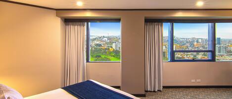 Ein Hotelzimmer mit einem großen Bett, zwei mit Vorhängen versehenen Fenstern, Blick auf die Stadt und Teppichboden.