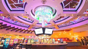 Interior de um cassino com um lustre central, várias máquinas de jogo e uma área de bar.