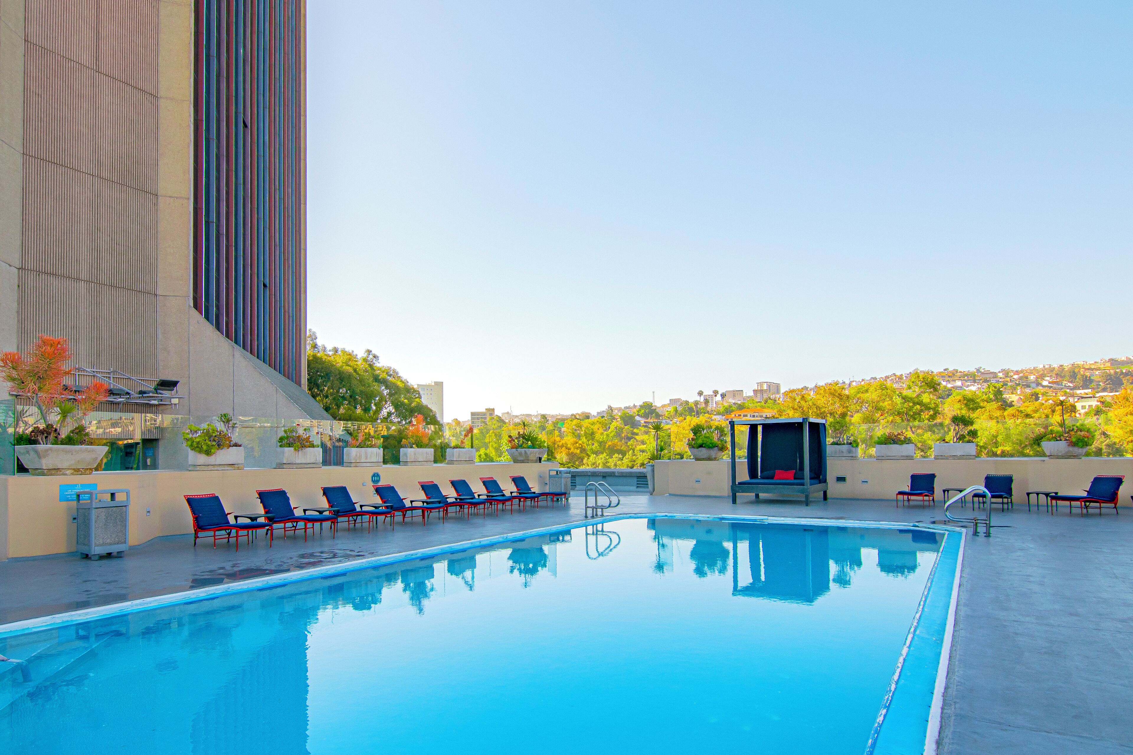A rooftop swimming pool with blue chairs, a small seating area, and a view of a cityscape.