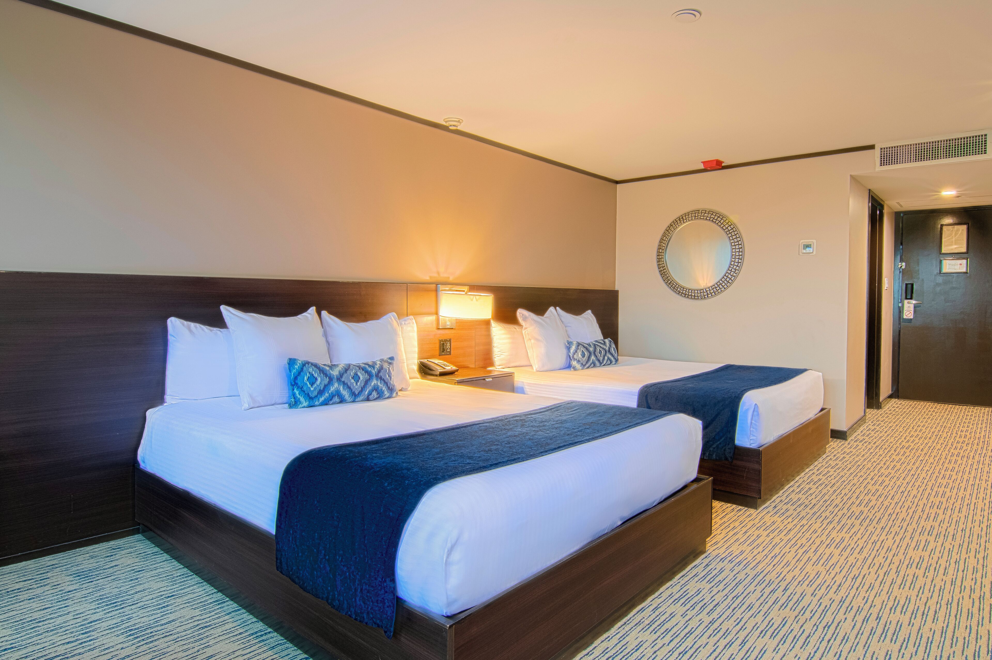 Une chambre d’hôtel avec deux lits, une table de chevet, une lampe, un miroir et un sol recouvert de moquette.
