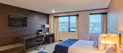 A hotel room with two beds, a desk, a TV, and a view of a cityscape.