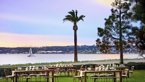 An outdoor dining area with tables and chairs arranged on a grassy lawn, overlooking a body of water with a sailboat in the distance. - Sheraton San Diego Resort (San Diego)