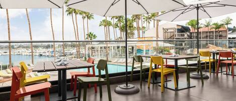 An outdoor dining area with tables and chairs, umbrellas, and a view of a marina with boats.