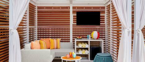A modern outdoor lounge area with a grey sofa, colorful cushions, a small round table with drinks, and a teal stool.