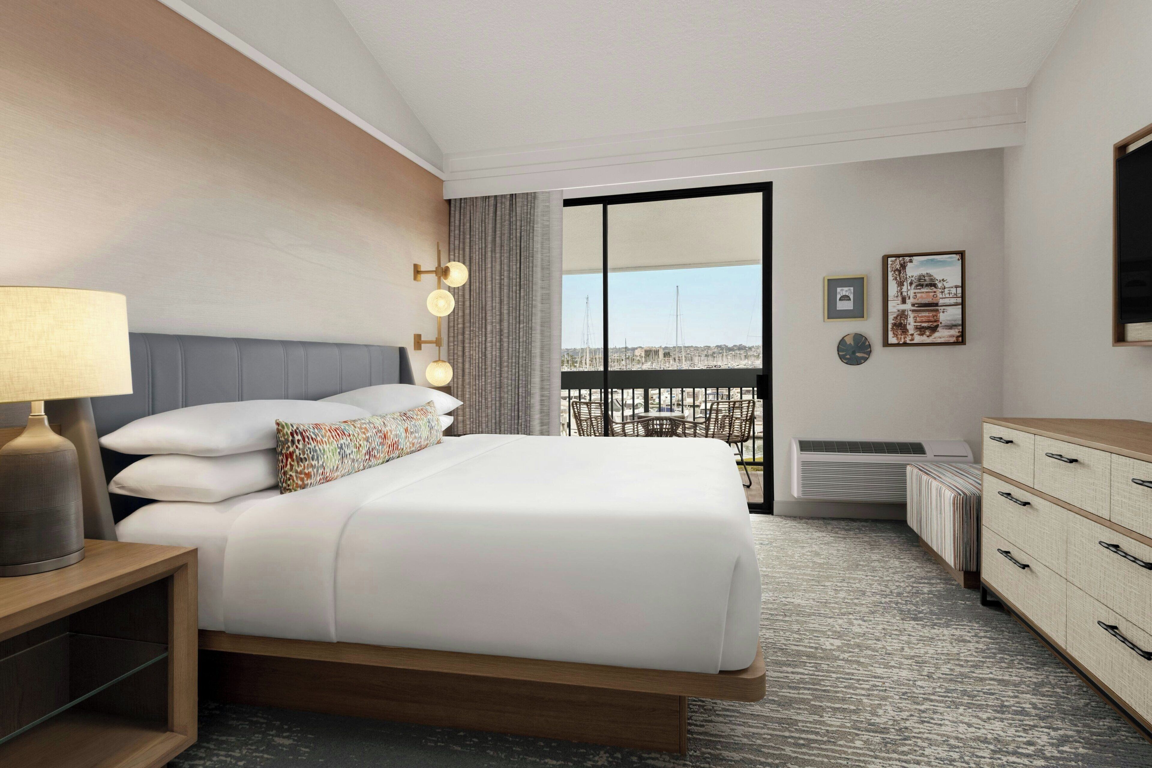 A hotel room with a large bed, a bedside table with a lamp, a dresser, and a view of the city through a balcony.