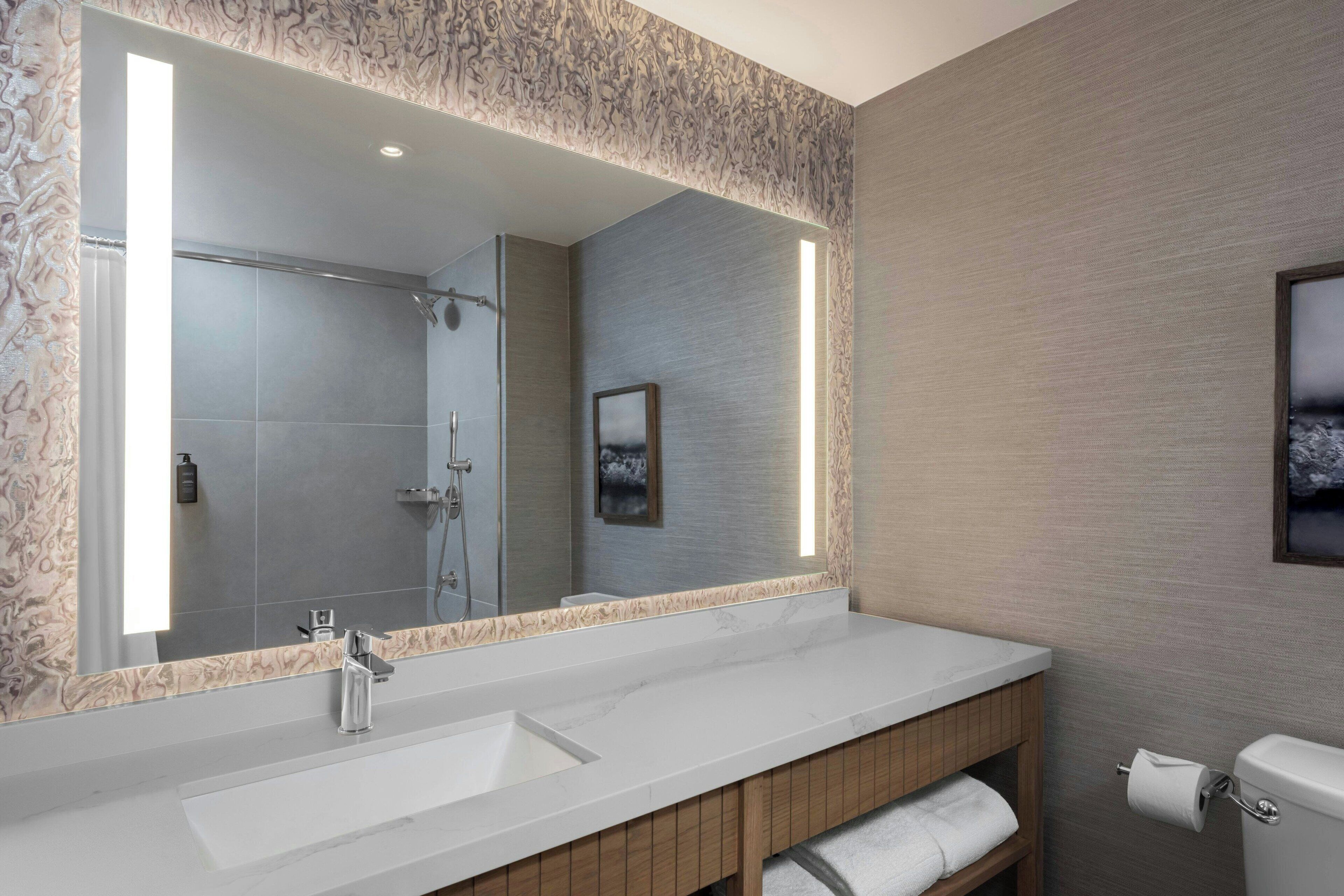 A modern bathroom with a large mirror, a white sink, a shower, and a toilet.