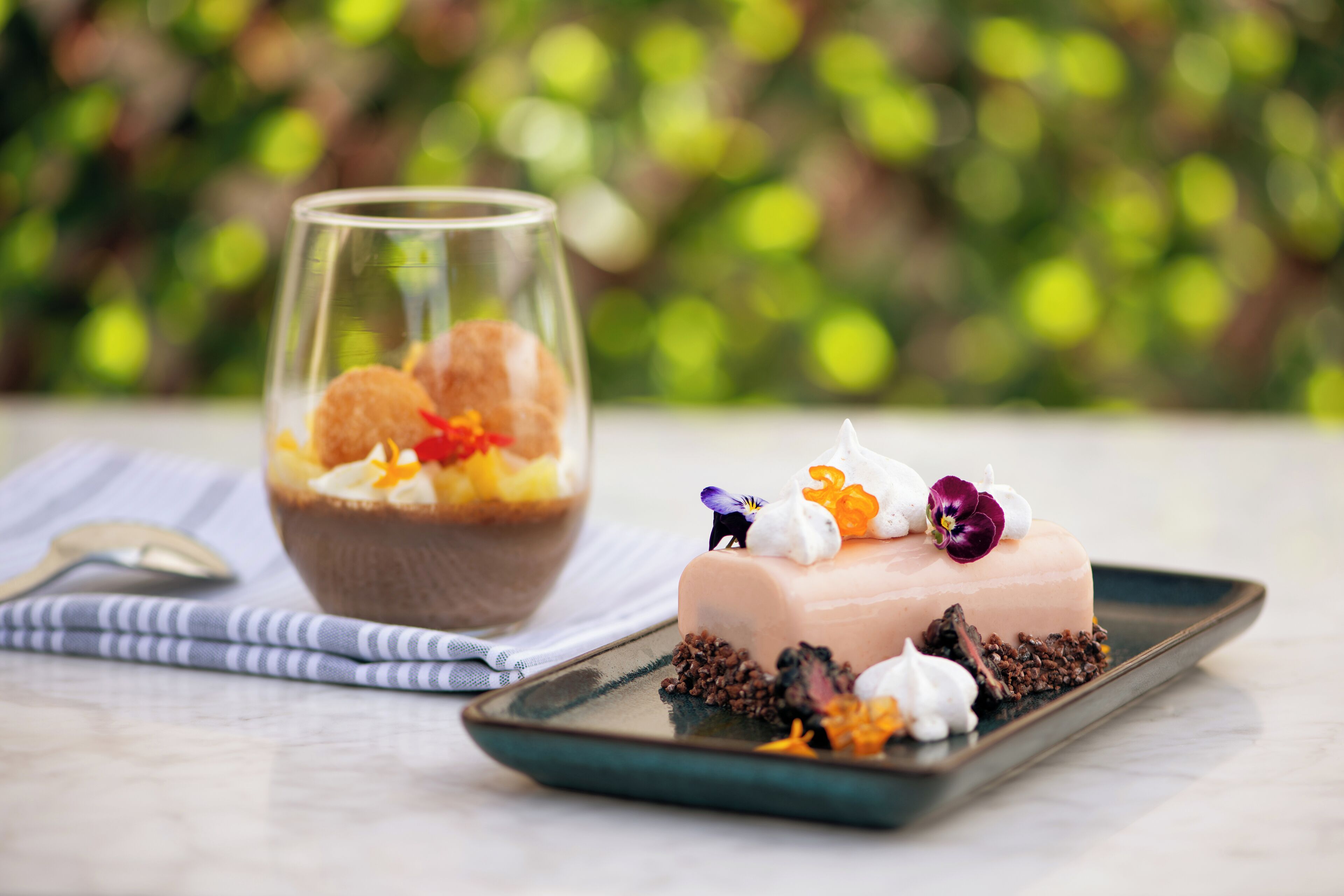 Un dessert accompagné d’un verre de ce qui semble être une boisson à base de fruits, d’une petite figurine et de quelques fleurs décoratives disposées sur une assiette.
