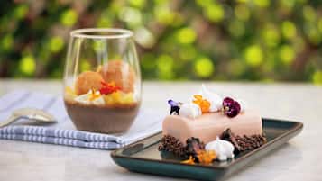 Un dessert accompagné d’un verre de ce qui semble être une boisson à base de fruits, d’une petite figurine et de quelques fleurs décoratives disposées sur une assiette.
