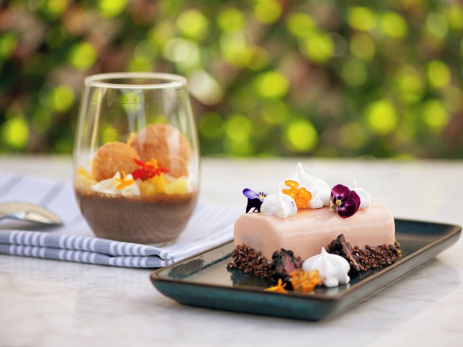 Un dessert accompagné d’un verre de ce qui semble être une boisson à base de fruits, d’une petite figurine et de quelques fleurs décoratives disposées sur une assiette.