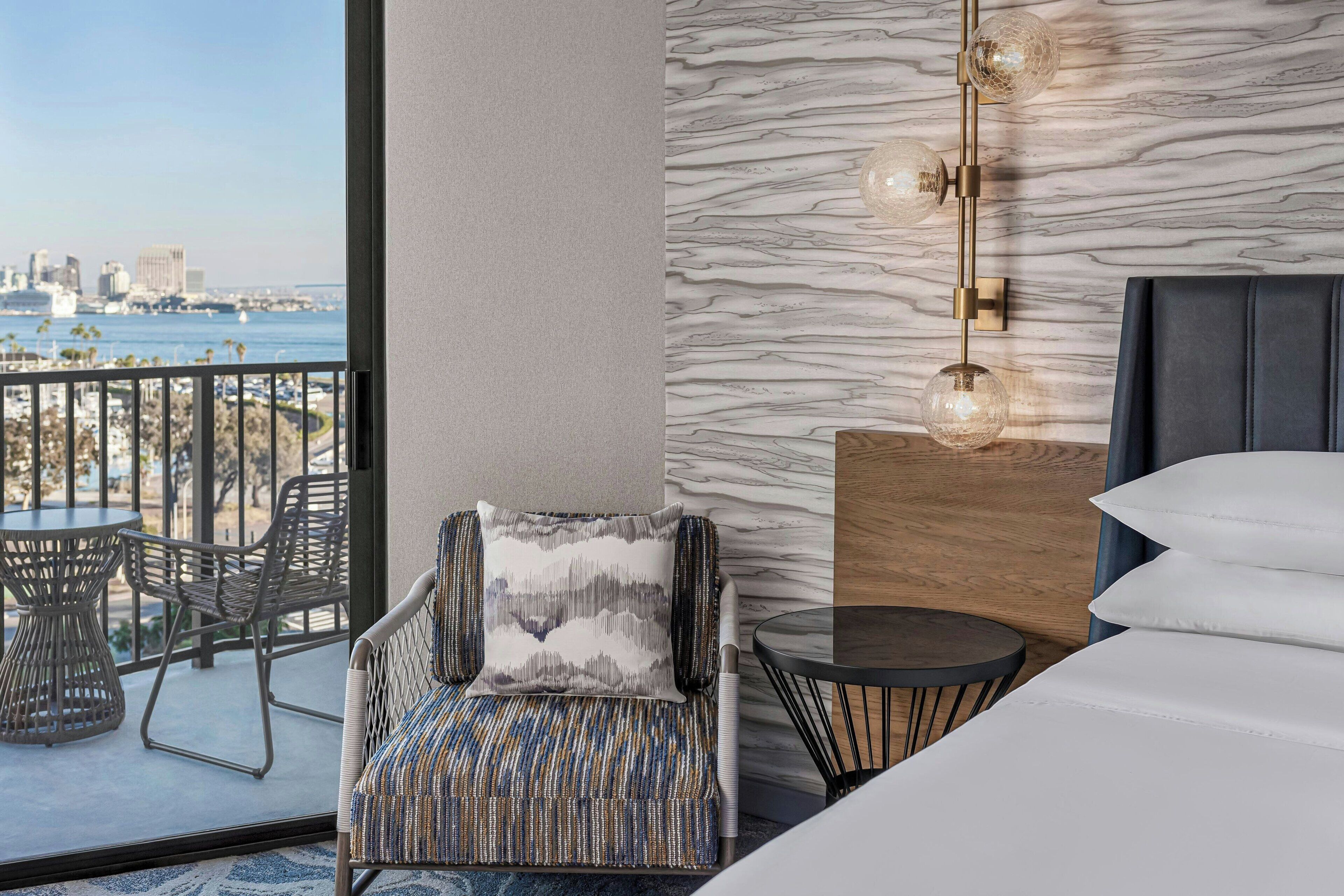 A modern hotel room with a balcony, a city view, a marble-patterned wall, and a stylish bed.