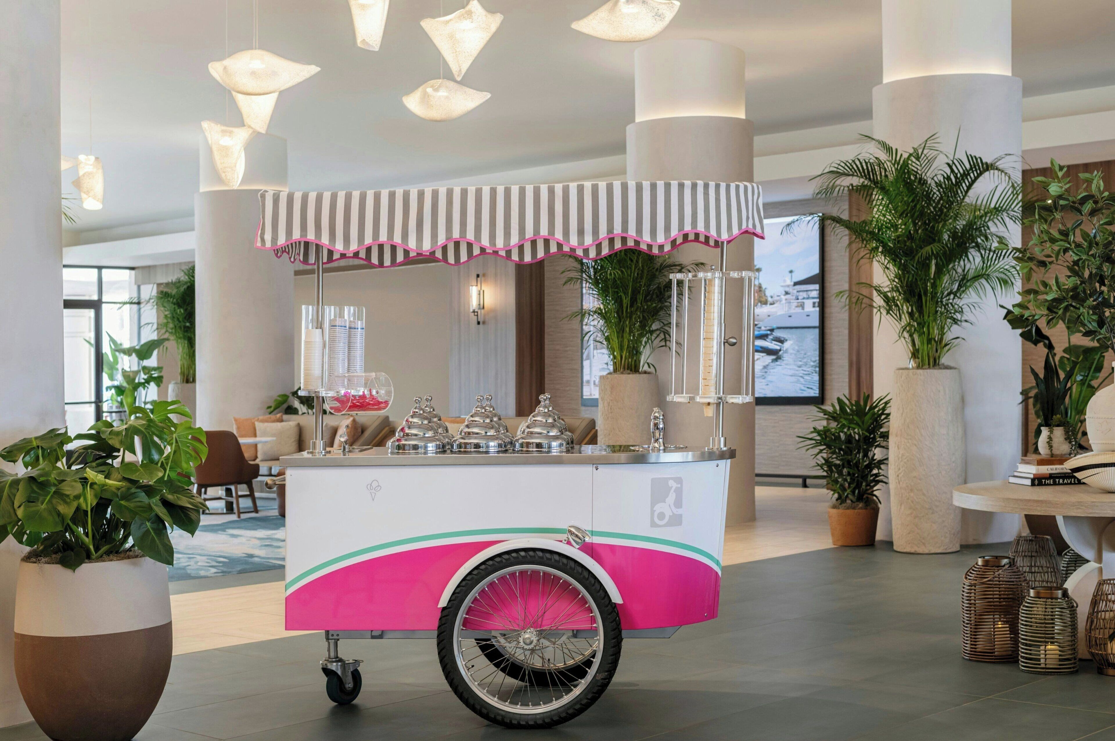 Een roze en witte foodtruck met een gestreept luifeltje, gelegen in een ruime lobby met grote zuilen, potplanten en een zitgedeelte.