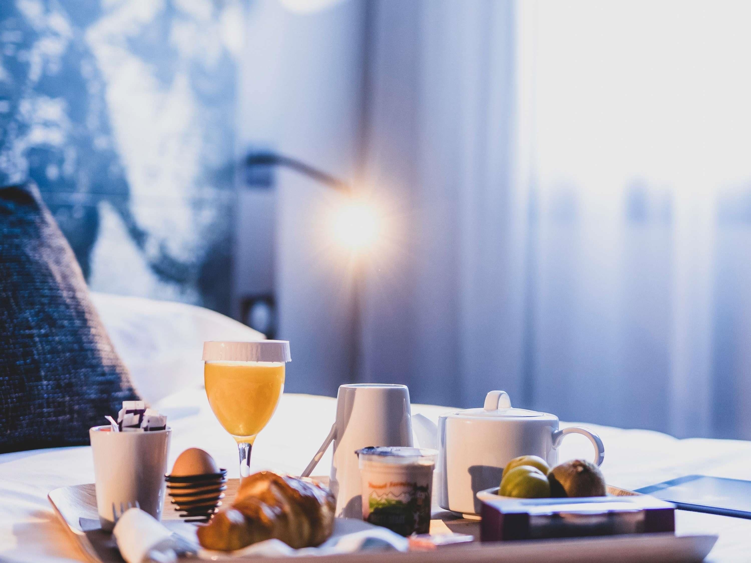A breakfast tray with croissants, a glass of orange juice, a mug, and a teapot on a bed with a view of trees and a cityscape.