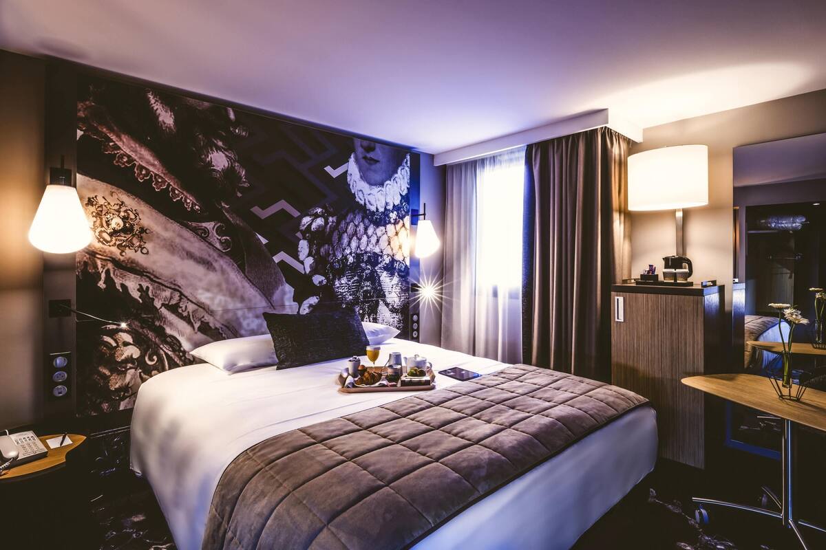 Quarto de hotel com uma cama grande, uma escrivaninha com abajur e um quadro na parede.