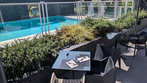 An outdoor pool area with a glass barrier, surrounded by greenery and a covered seating area with tables and chairs. - Mercure Pau Palais Sports (Pau)