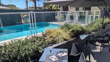 An outdoor pool area with a glass barrier, surrounded by greenery and a covered seating area with tables and chairs.