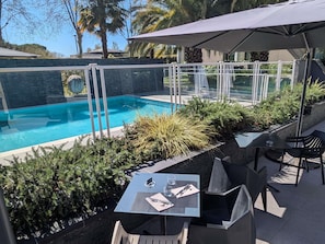 Un espace piscine extérieur doté d’une barrière en verre, entouré de verdure et comprenant un espace de repos couvert avec des tables et des chaises.