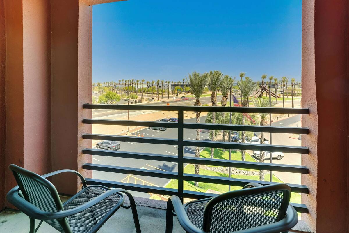 A balcony with metal railings and two chairs, overlooking a street with palm trees and a playground.