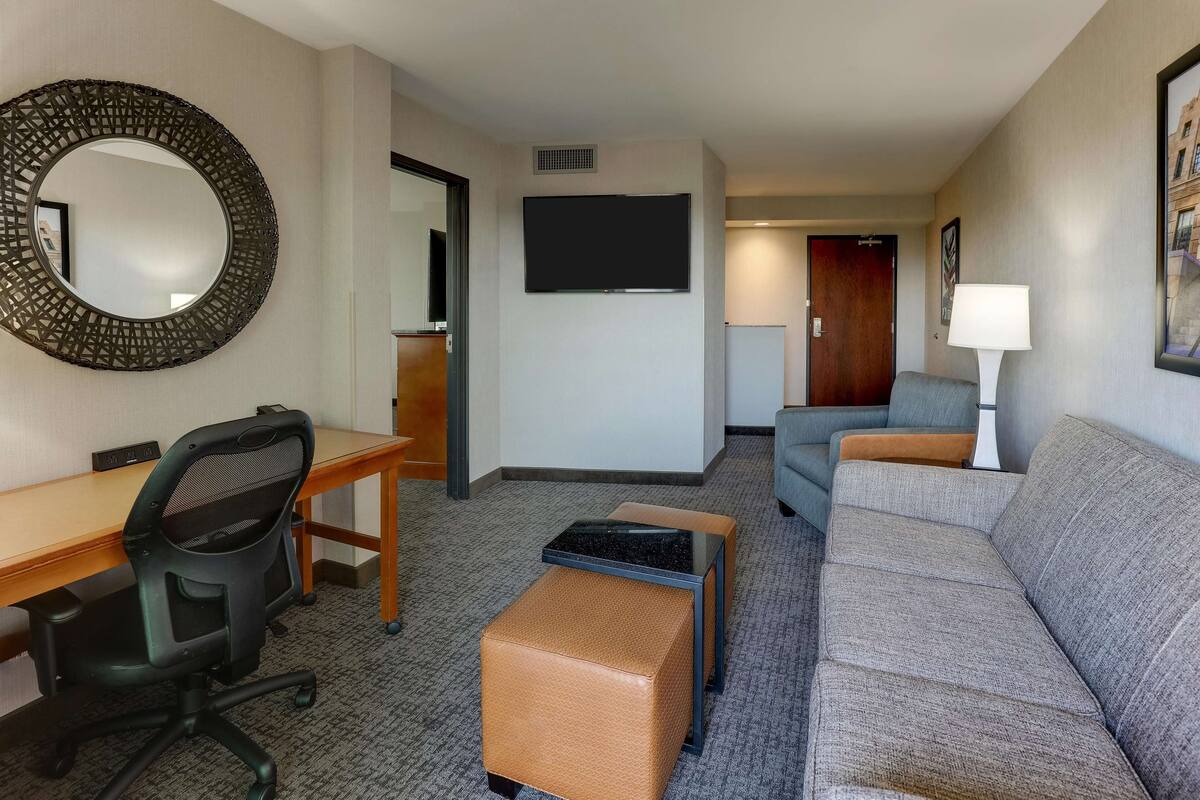 A hotel room with a desk, office chair, sofa, ottoman, and a TV mounted on the wall.