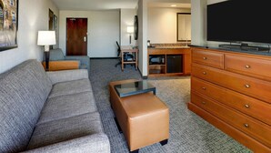 A hotel room with a sofa, ottoman, wooden dresser, flat-screen TV, desk with chair, lamp, and a framed picture on the wall. - Drury Inn & Suites Phoenix Airport (Phoenix)