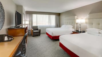 A hotel room with two beds, a desk with a television, a chair, and a large window with curtains.