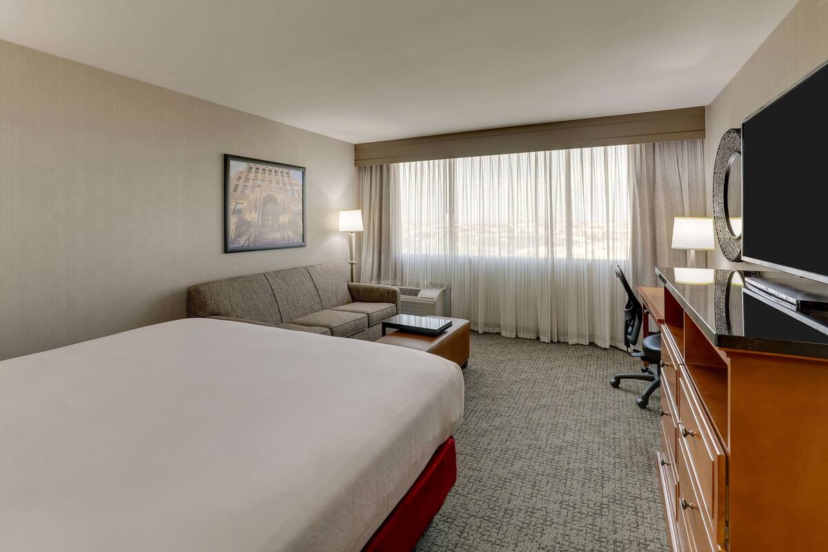 A hotel room with a large bed, a sofa, a desk with a computer, a television, and a window with curtains.