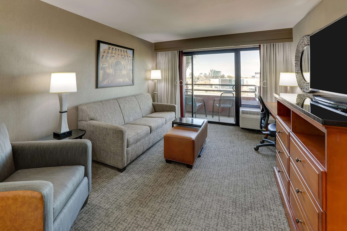 A hotel room with a sofa, ottoman, TV, and a view through the glass door.