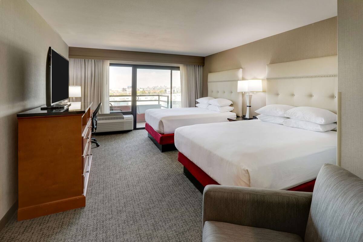 A hotel room with two beds, a TV, a desk, and a view of the city.
