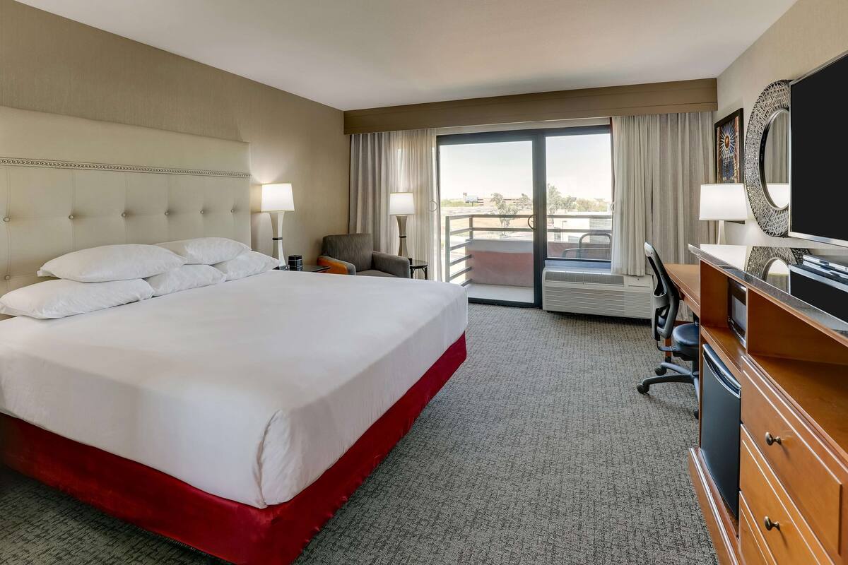 A hotel room with a large bed, a desk, a chair, a TV, and a balcony with a view.