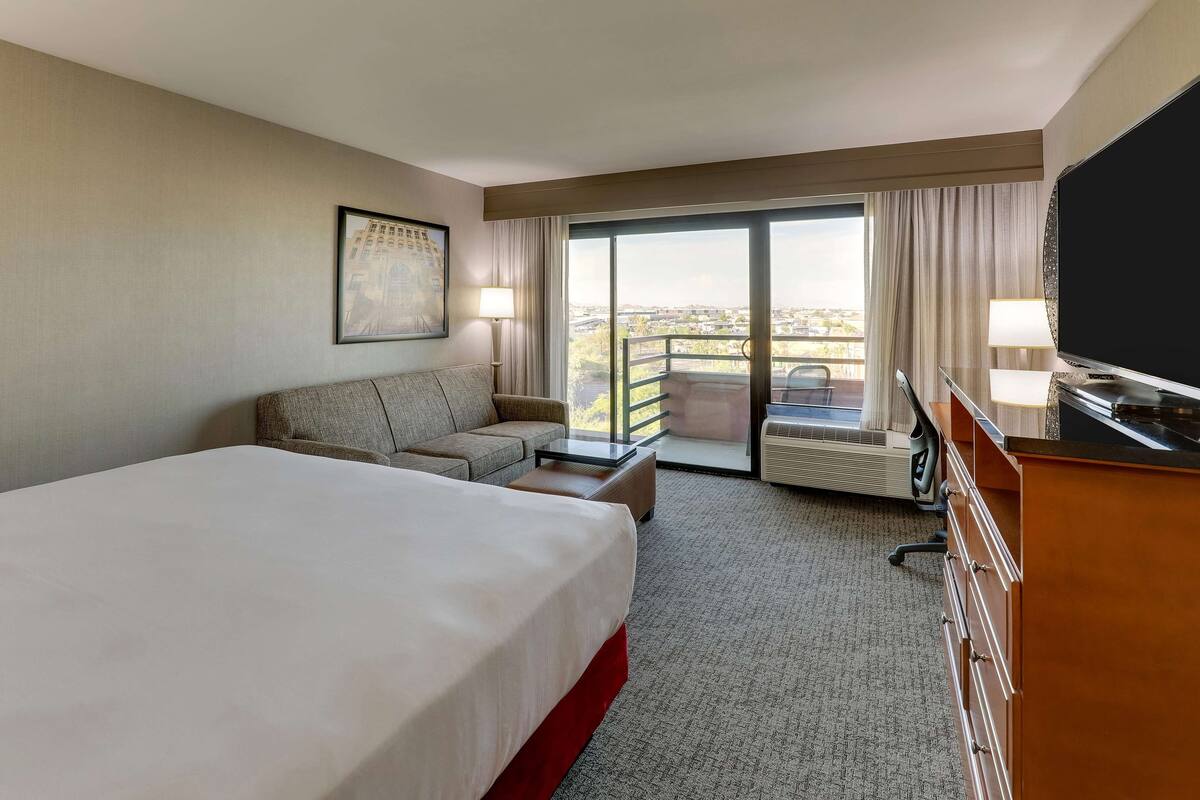 A hotel room with a large bed, a sofa, a desk, a TV, and a balcony with a view.