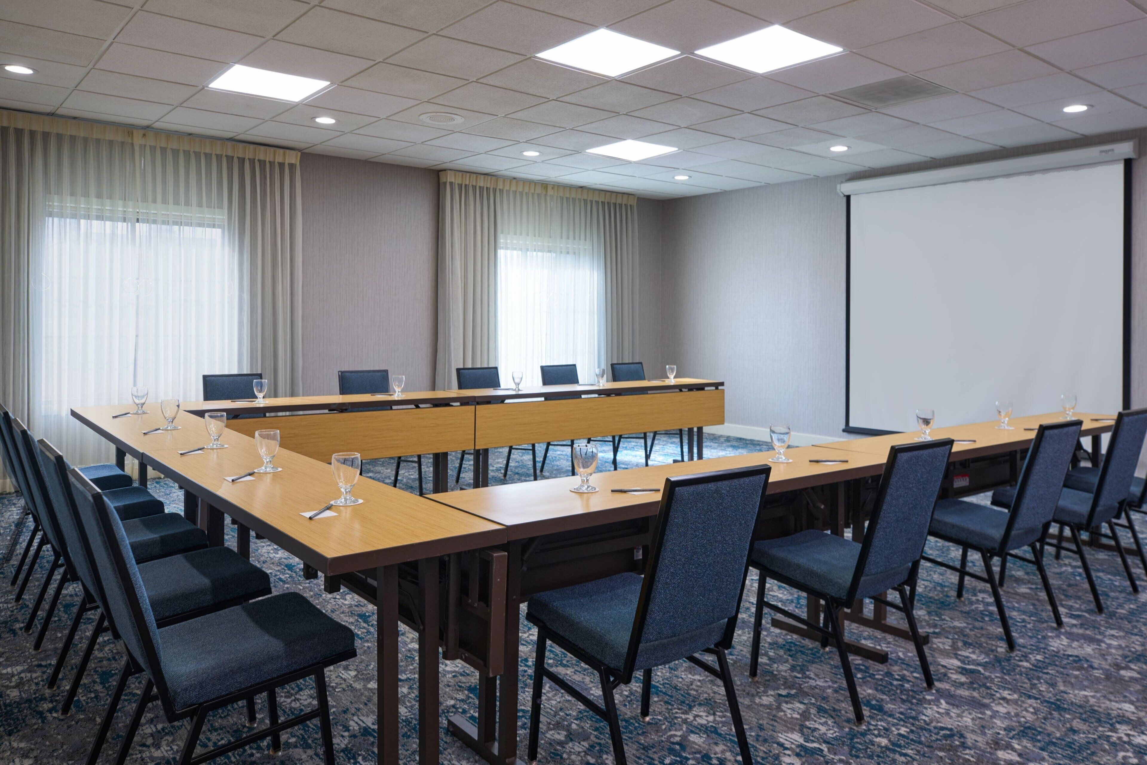 Un salón de conferencias con una larga mesa, sillas, una pantalla de proyector y un amplio ventanal con cortinas.