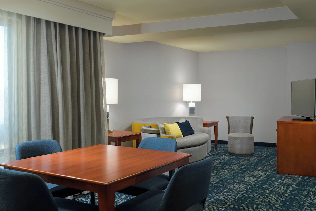 A hotel room with a wooden table, blue chairs, a grey sofa with yellow and blue cushions, a grey armchair, a small round ottoman, and a television on a wooden cabinet.