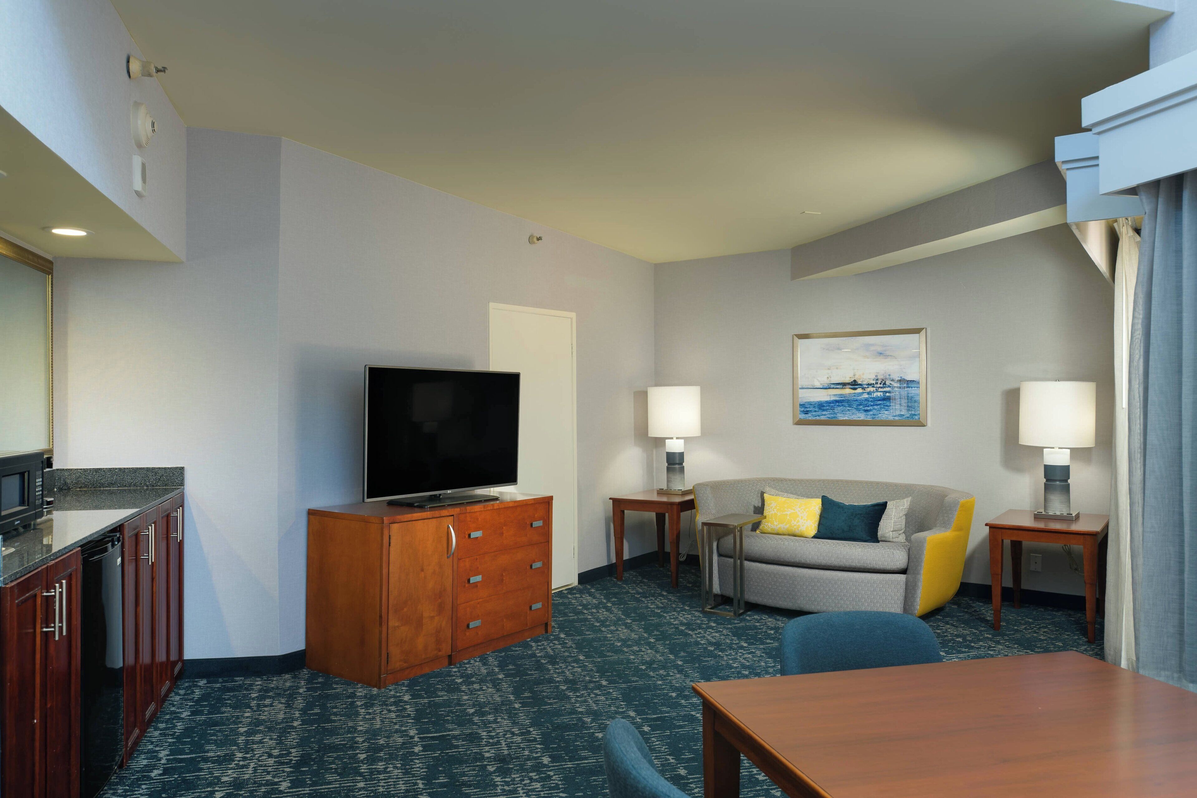 A hotel room with a flat-screen TV, a sofa, two bedside lamps, a dining table, and a kitchen area with a microwave.