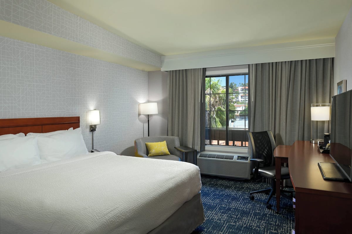 A hotel room with a large bed, two bedside lamps, a desk with a chair, an air conditioning unit, and a window with curtains.