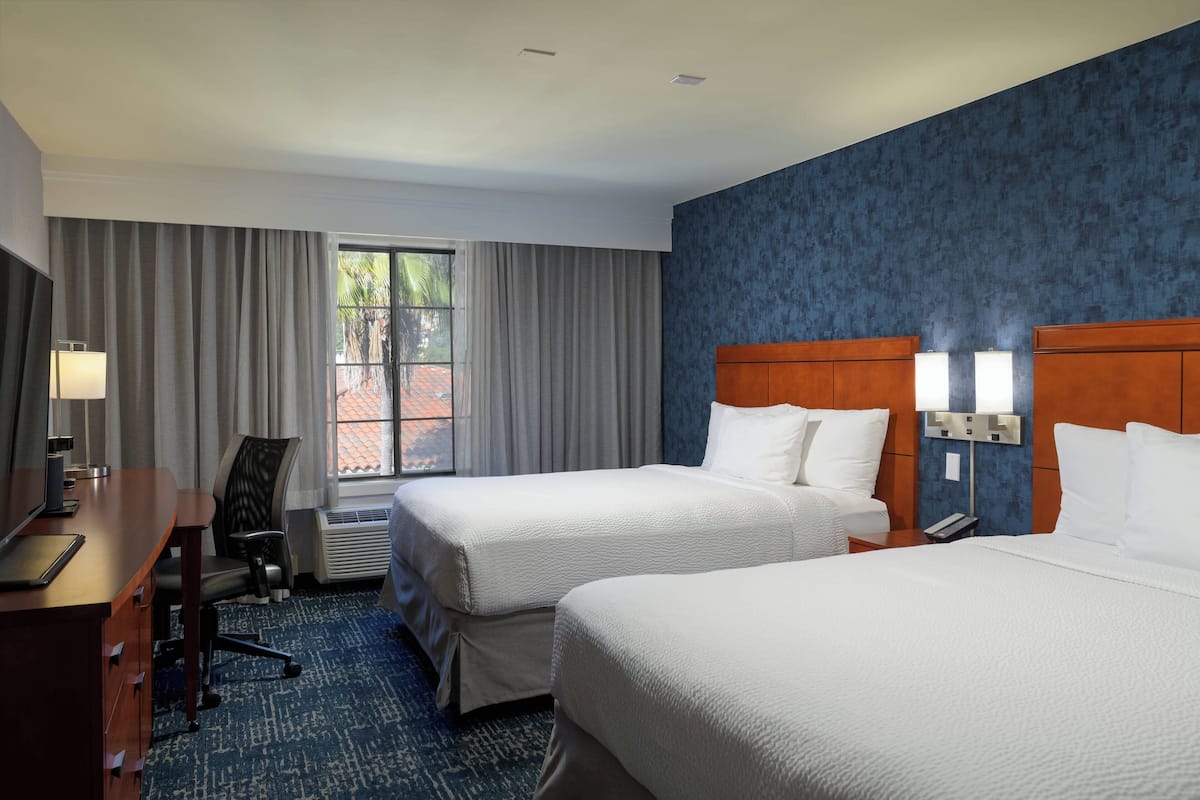 A hotel room with two beds, a desk, a chair, a lamp, and a window with curtains.