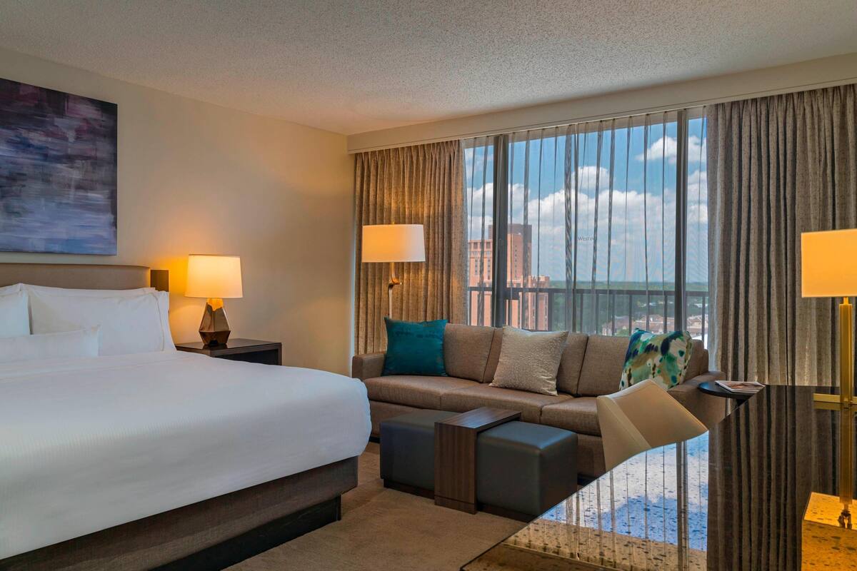 Habitación de hotel con una cama grande, un sofá, una mesita y vistas a la ciudad desde la ventana.