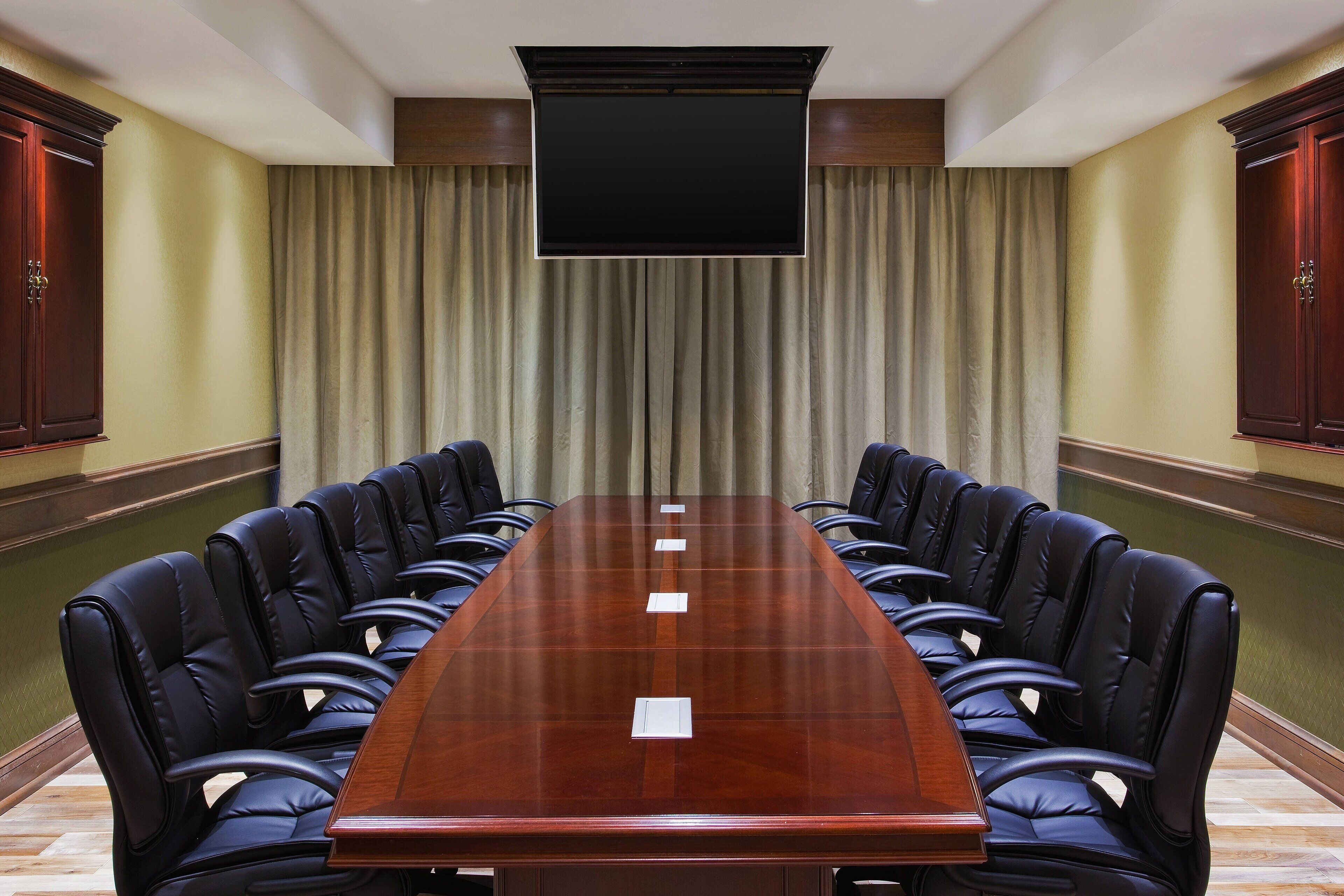 A conference room with a long table, black leather chairs, a flat-screen TV, and a large window with curtains.