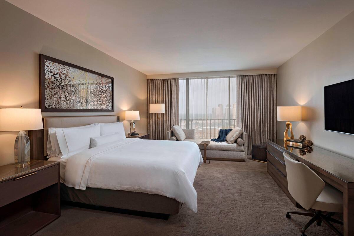 Una habitación de hotel con una cama grande, dos mesitas de noche con lámparas, un escritorio con silla, un sofá y un ventanal con cortinas.