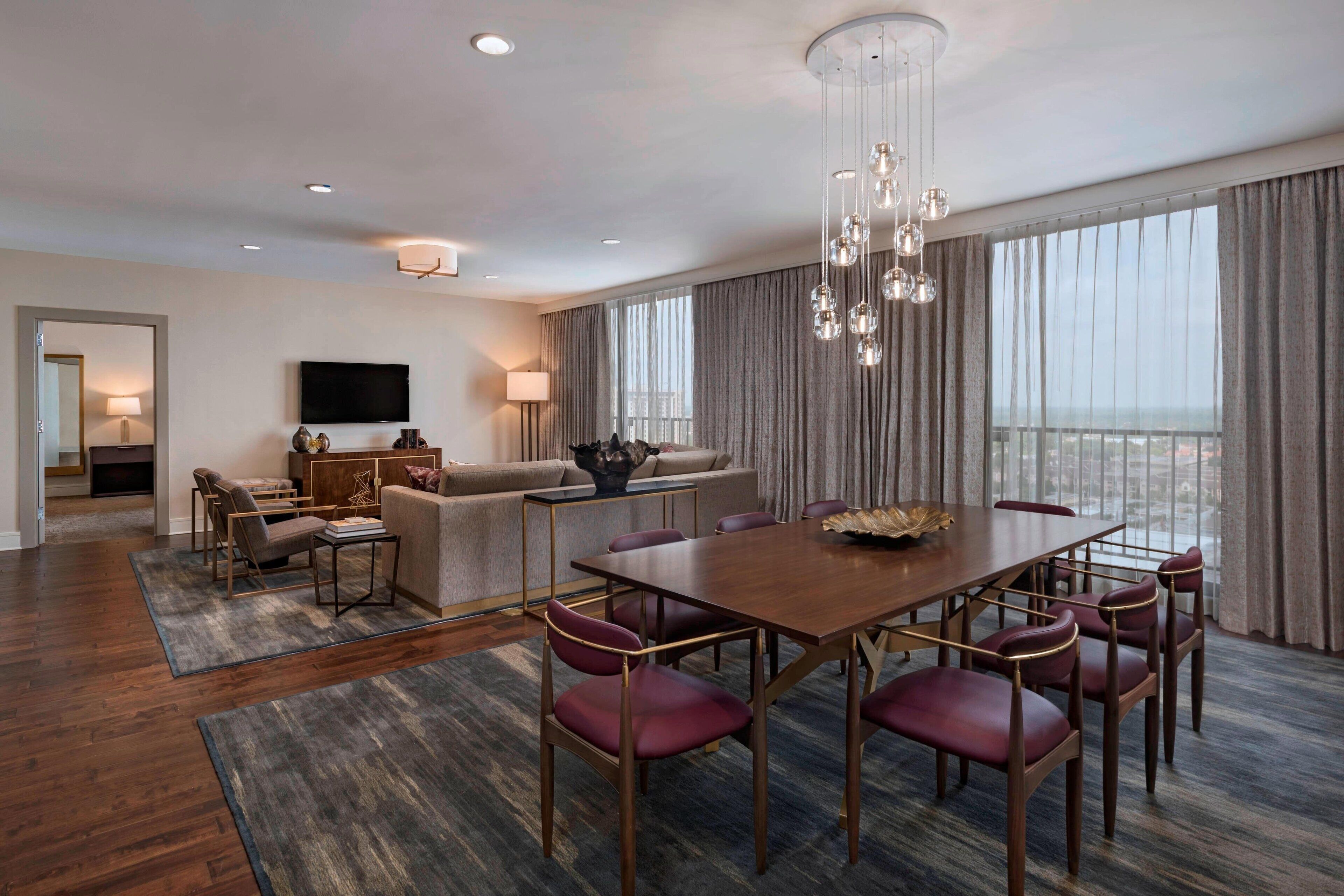 A modern hotel room with a dining area, a sofa set, a flat-screen TV, and a large window with curtains.