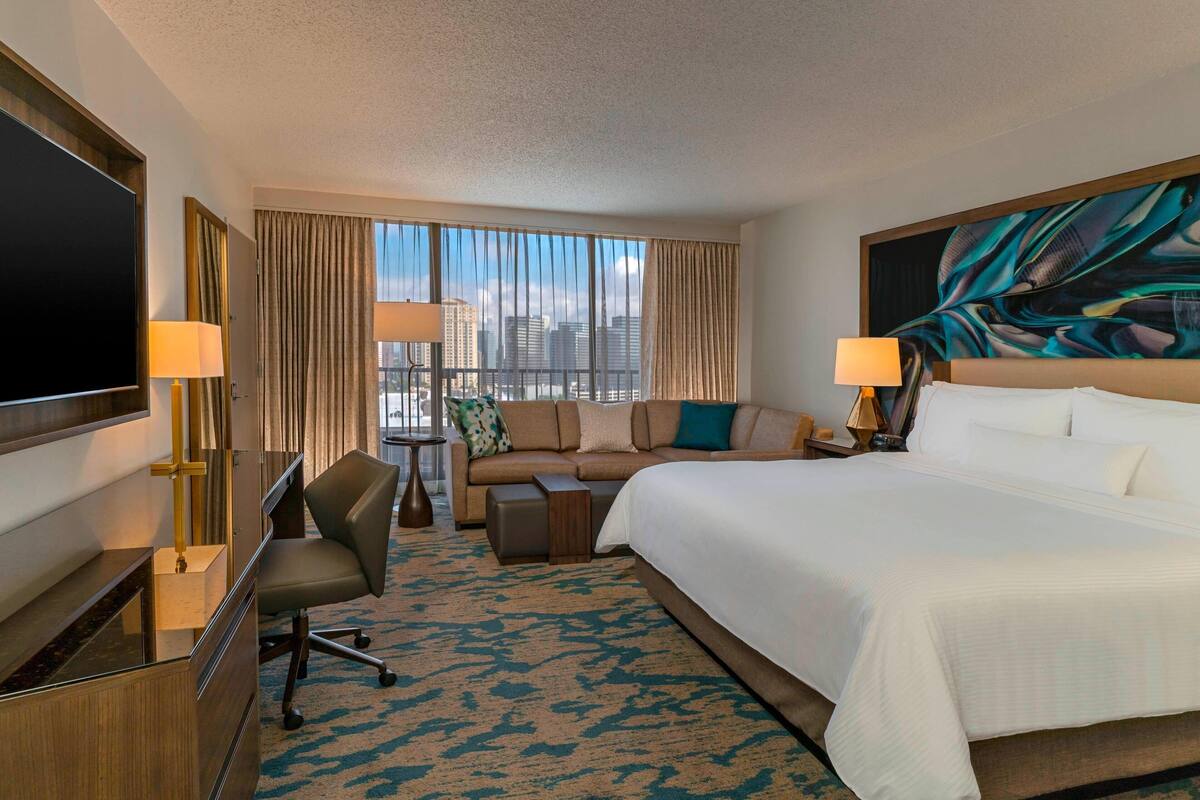 Habitación de hotel con una cama grande, un sofá, un escritorio con televisor y vista a la ciudad desde la ventana.