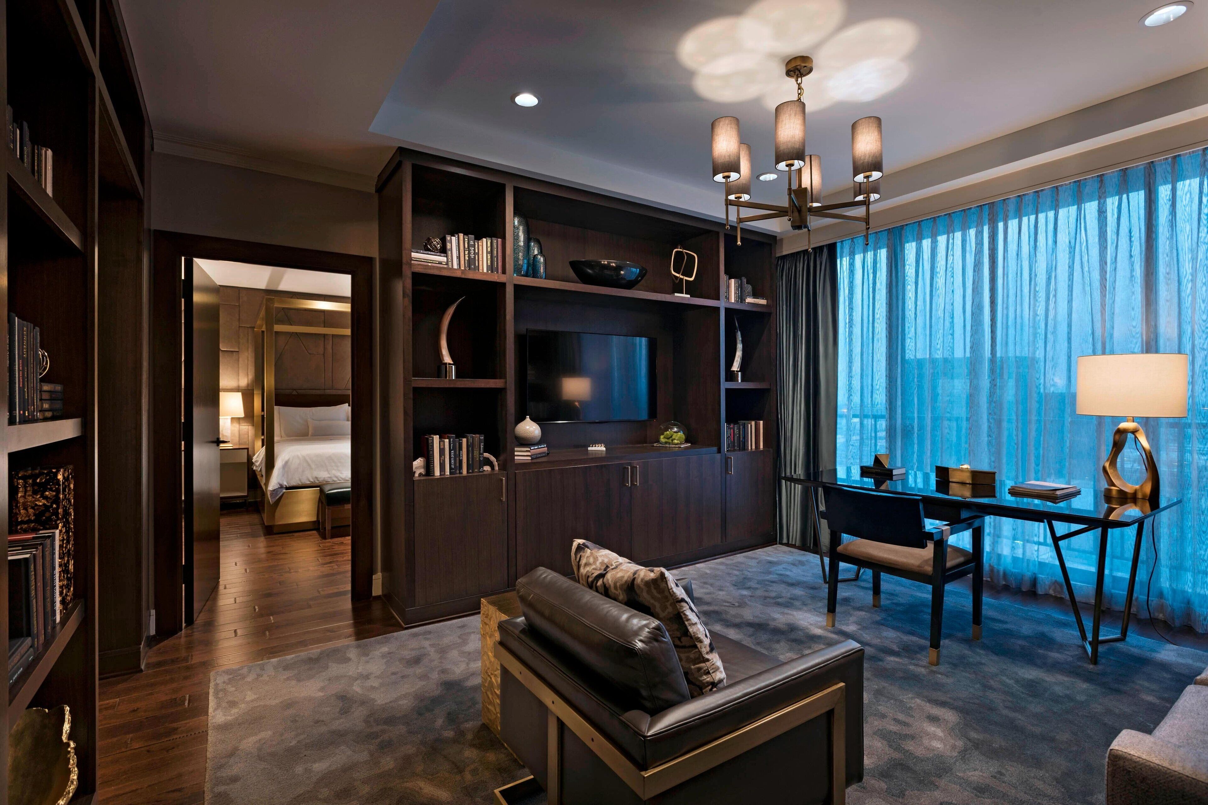 A modern hotel room with a large bed, a seating area with a sofa and armchair, a dining table, a TV, and a bookshelf.