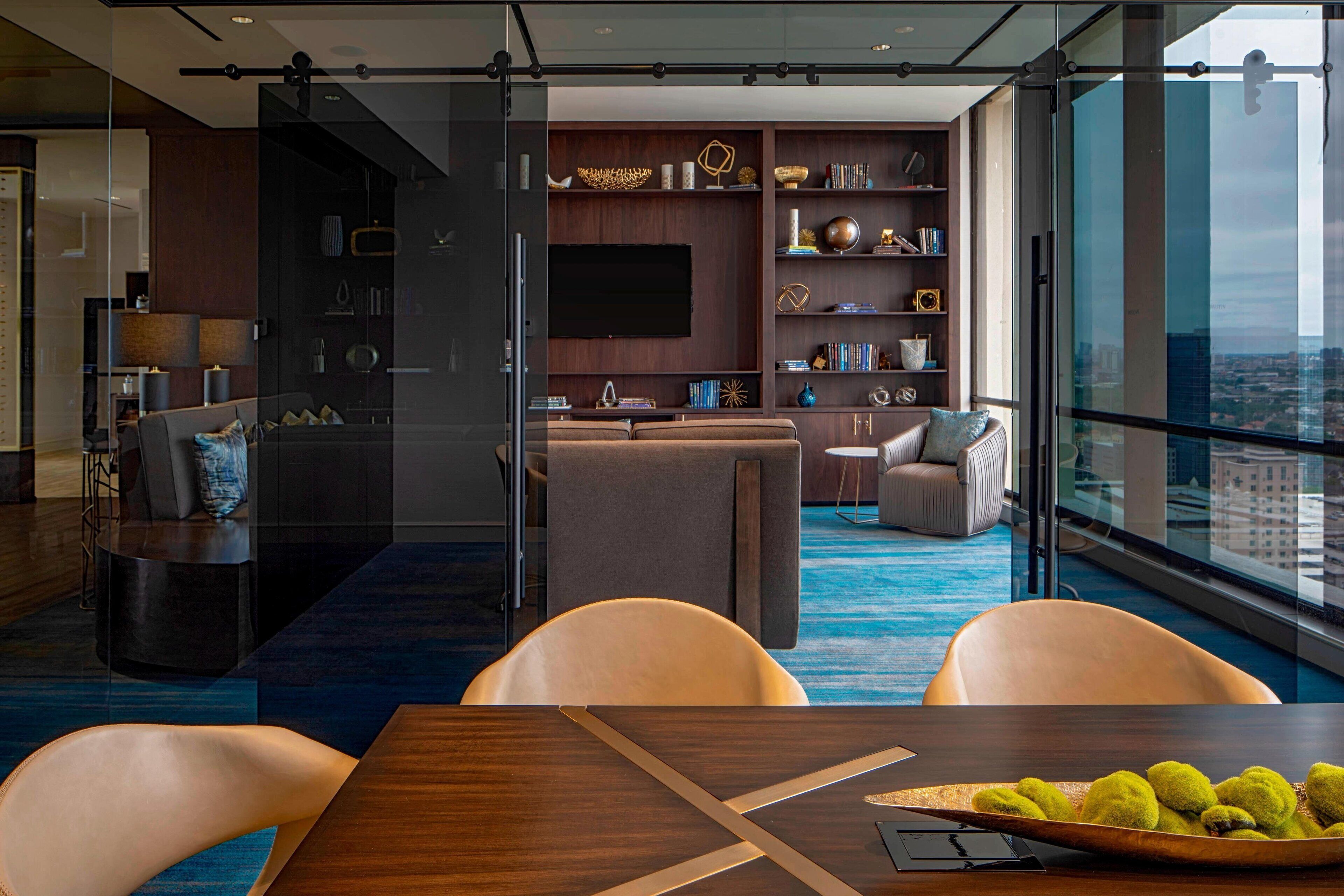 A modern hotel room with a large window, a dining area with a wooden table, and a seating area with a sofa and chairs.