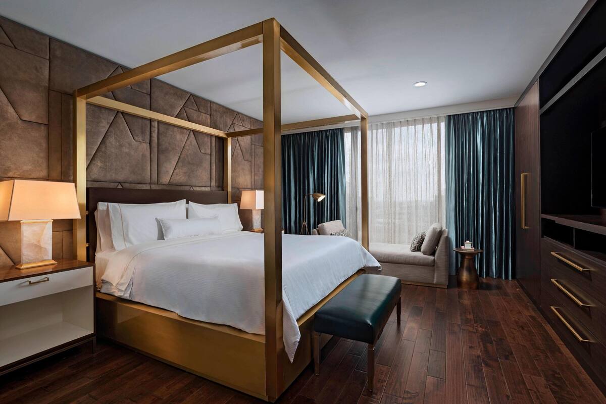Una cama con dosel de madera, una mesita de noche con lámpara, un banco, un sofá y un televisor a la derecha.