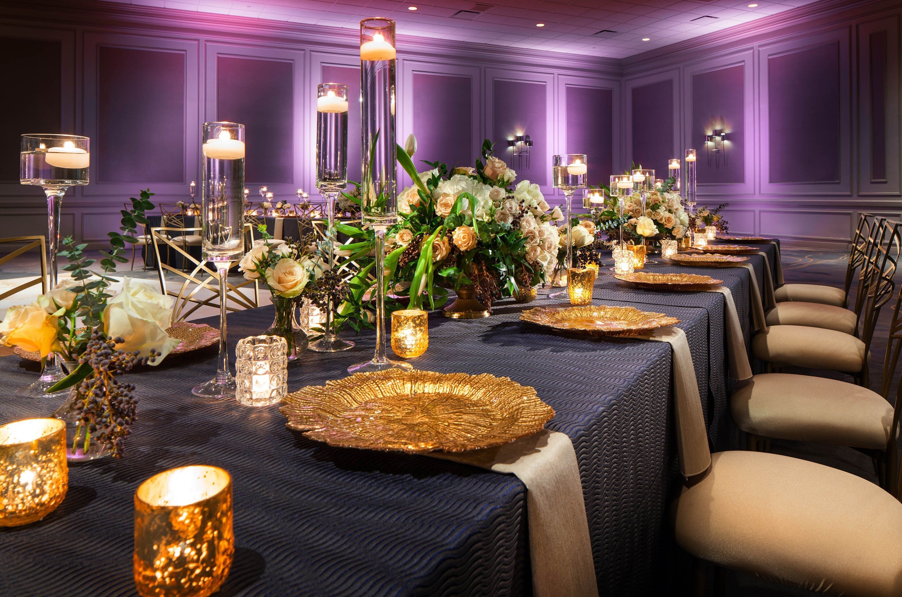 A banquet hall with long tables set for a formal event, adorned with floral arrangements, candles, and gold-rimmed plates.