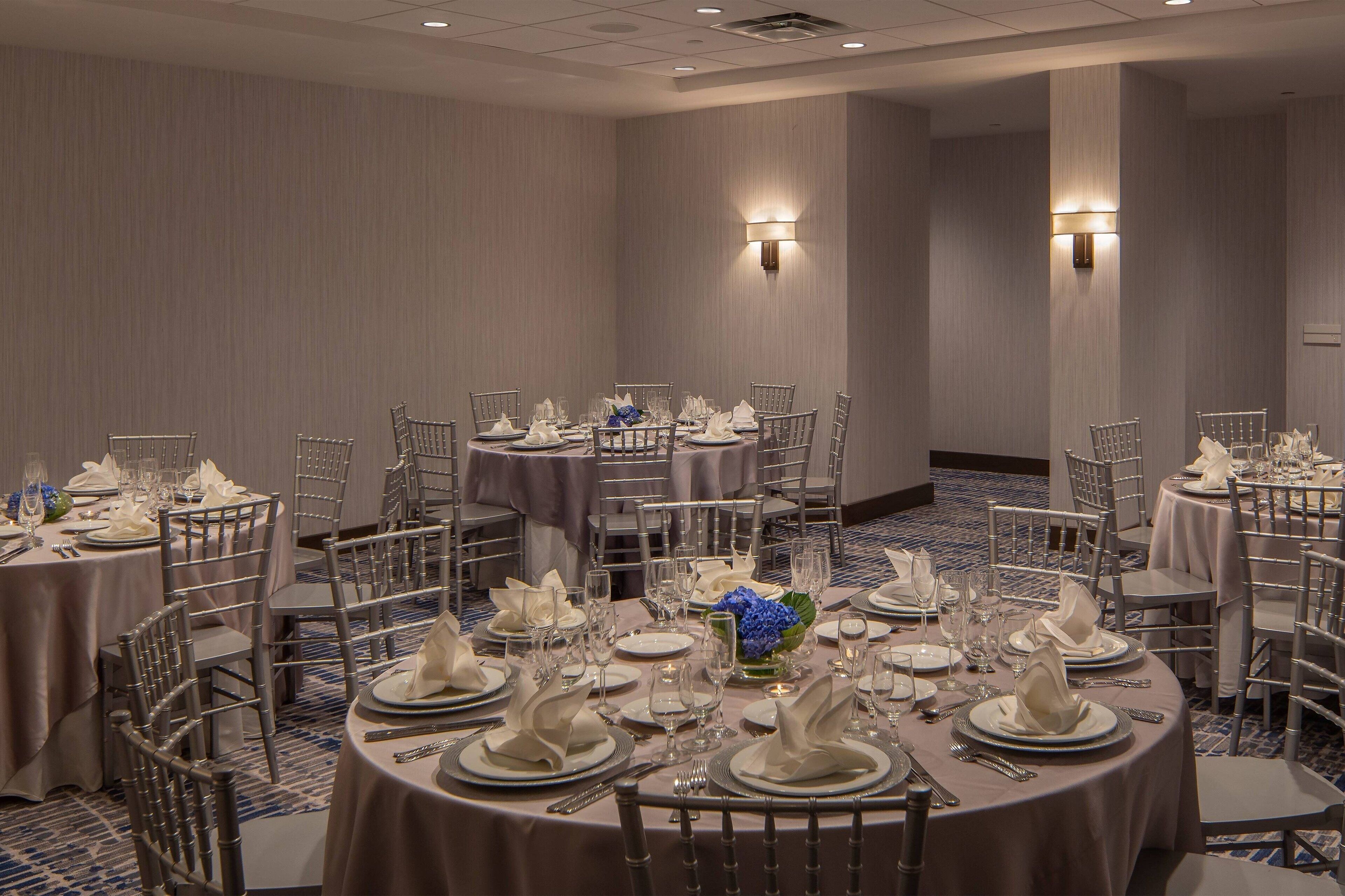 A banquet hall with round tables set for a formal event, featuring white tablecloths, silver chairs, and floral centerpieces.