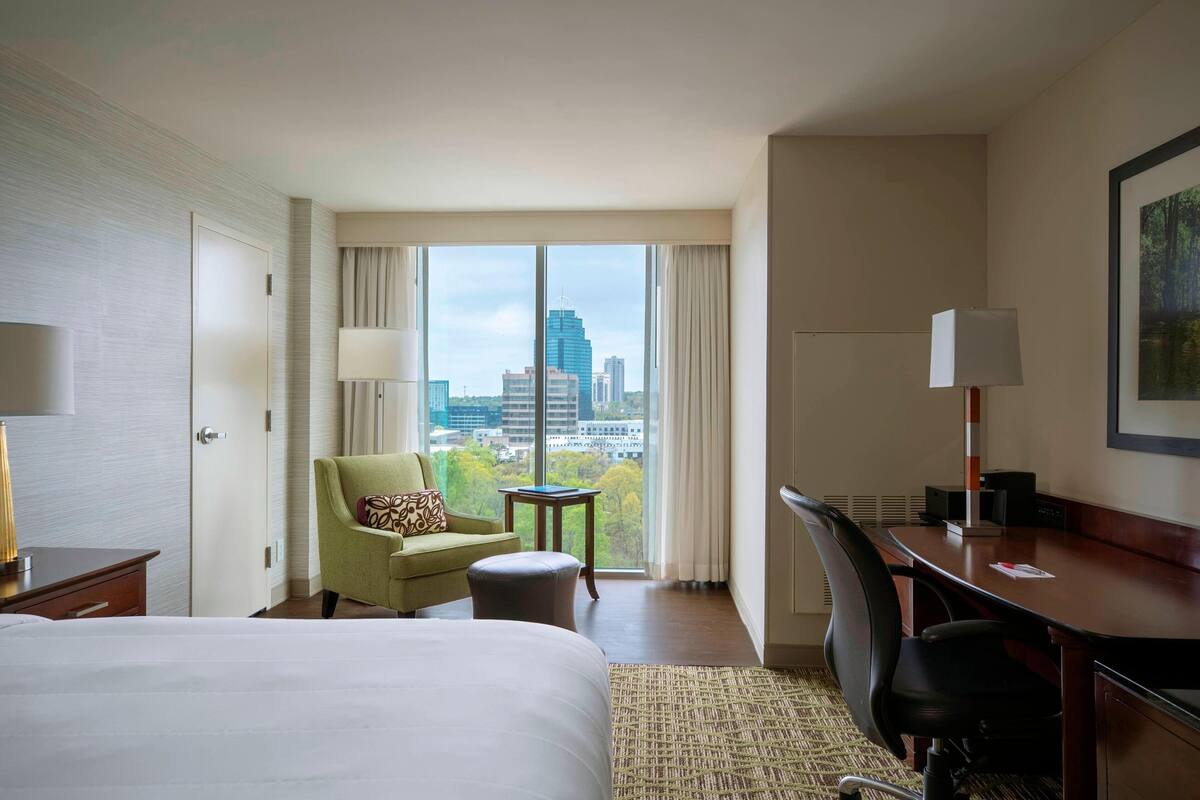 Hotelkamer met een groot bed, een bureau, een stoel, een bijzettafeltje en uitzicht op de stad door grote ramen.