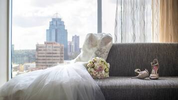 Una cama con un vestido de novia blanco y un ramo de flores, un sofá gris con un par de zapatos, y vista a la ciudad a través de una ventana.