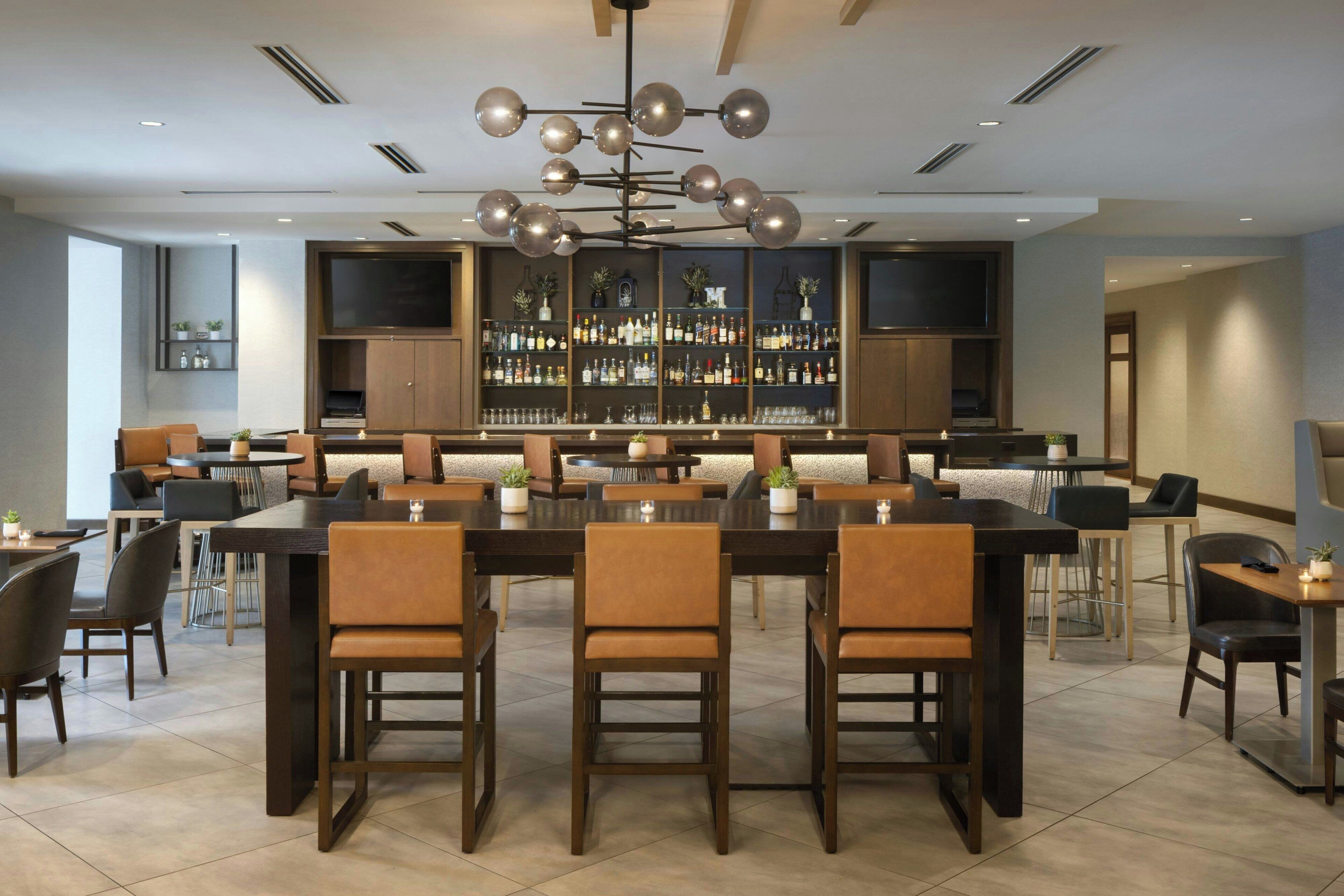 A modern bar area with a long counter, multiple seating options, and a large selection of bottles on shelves.