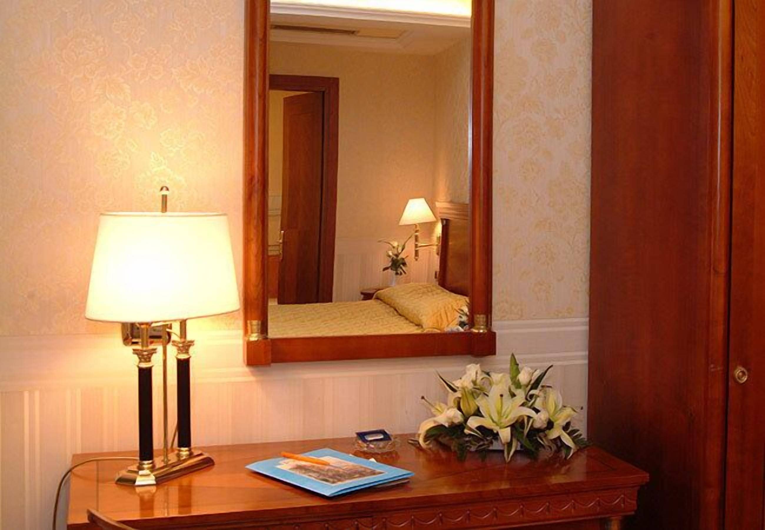A hotel room with a bedside table, a lamp, a mirror, and a floral arrangement.