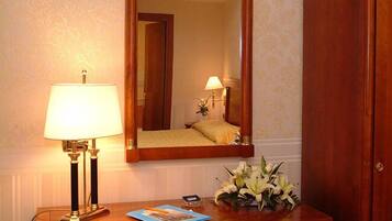 A hotel room with a bedside table, a lamp, a mirror, and a floral arrangement.